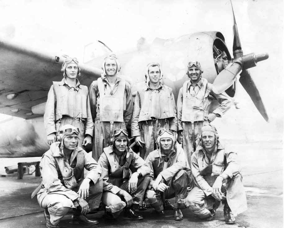 Eight Navy pilots in flight gear in front of a fighter